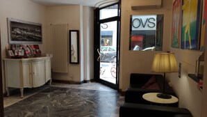 Un vestíbulo de hotel con piso de mármol, un mostrador de recepción, un cuadro enmarco con una vista urbana, un radiador y un ventanal con la señalización 'OVS'.
