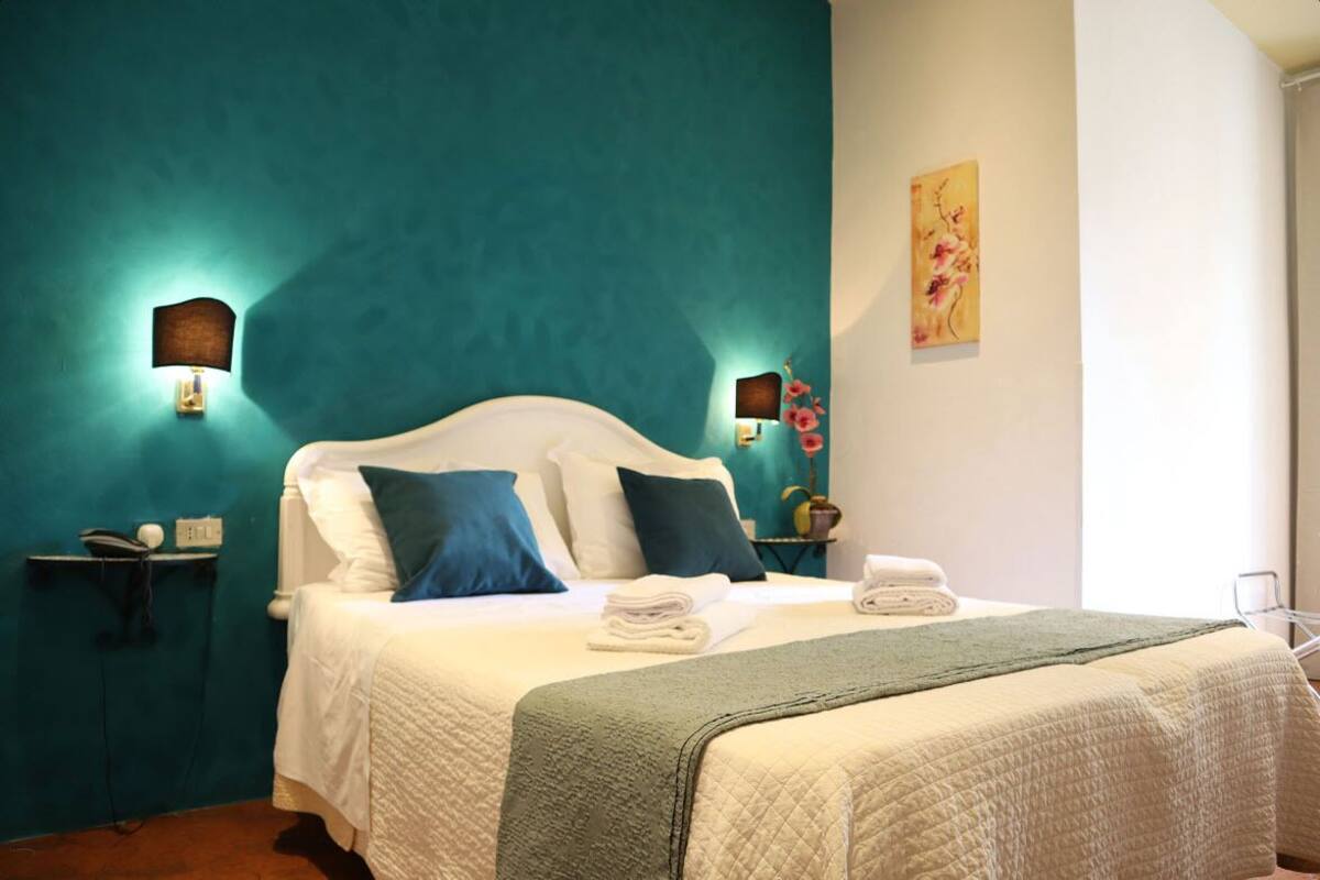 A hotel room with teal walls, a white bed with blue and white pillows, a green throw blanket, and two wall-mounted lamps.