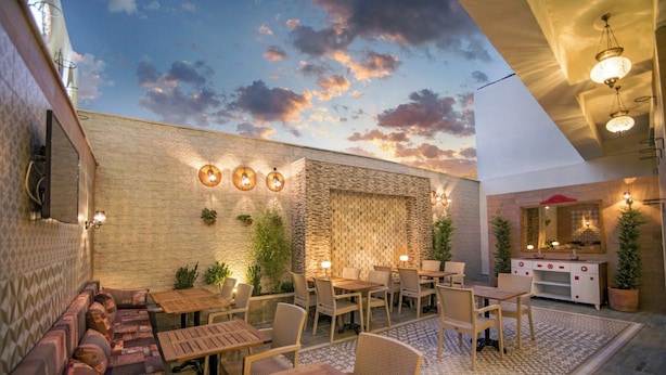 Un patio al aire libre con mesas y sillas de madera, una zona de estar con cojines, un muro de ladrillo y un aplique decorativo en el techo.