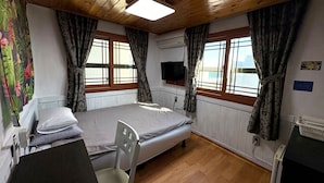 Une pièce comprenant un lit, un bureau et une chaise. Il y a deux fenêtres avec des rideaux, un téléviseur à écran plat fixé au mur et un radiateur posé sur le sol.