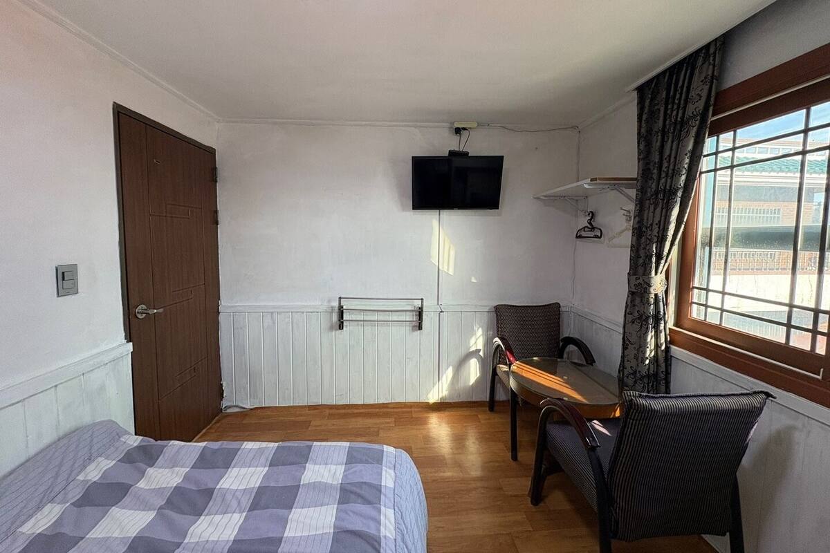 A room with two beds, a television mounted on the wall, a small table with a chair, and a window with curtains.