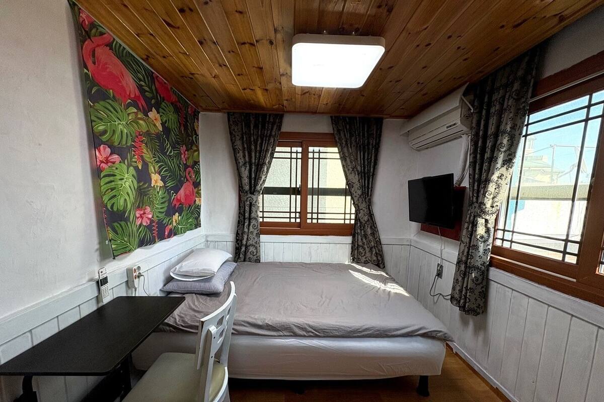 A bedroom with a bed, a desk, a chair, a TV, and a window with curtains.
