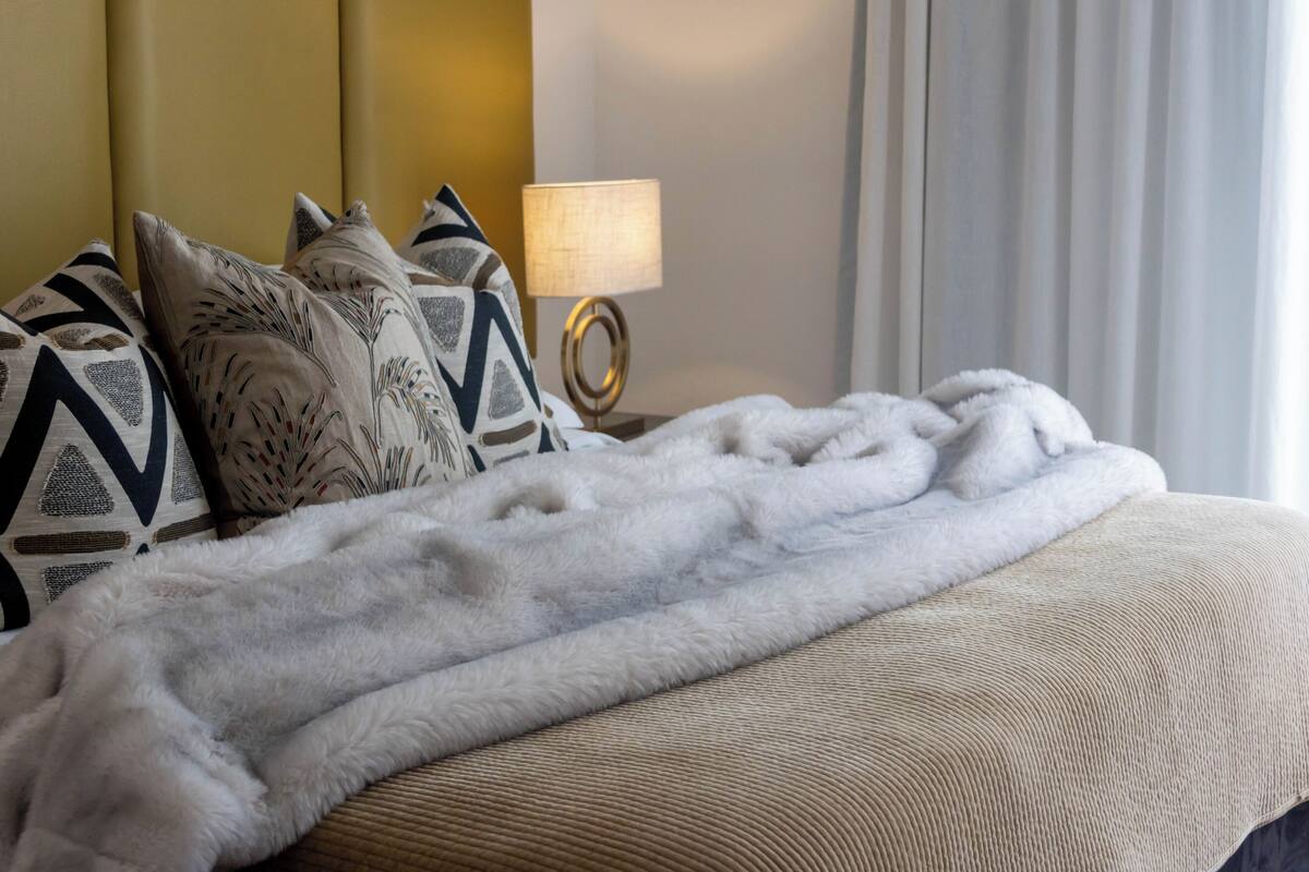 A neatly made bed with a white fluffy blanket, patterned pillows, and a bedside lamp.
