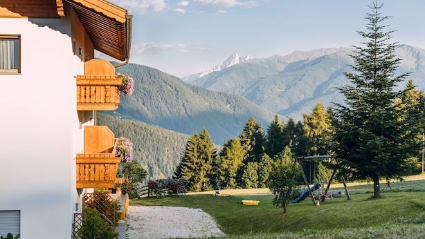 A mountain cabin with a wooden balcony, a garden with a swing set, and a view of distant mountains. - Residence Speckerhof (Rio di Pusteria)