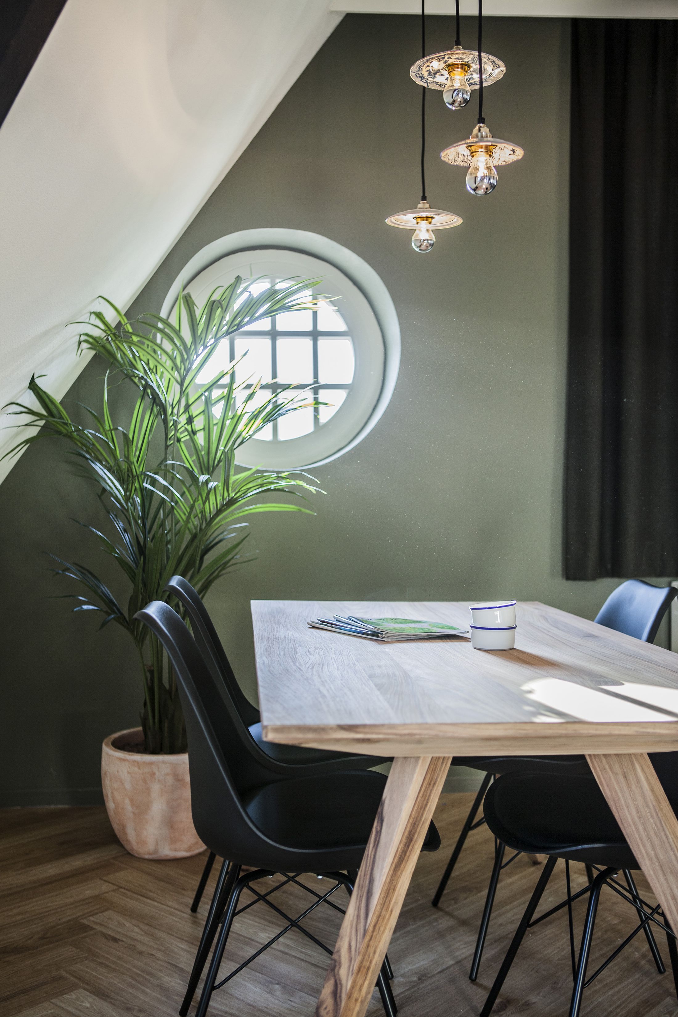 Uma sala de jantar com mesa de madeira, cadeiras pretas, uma planta em vaso e luminárias pendentes.