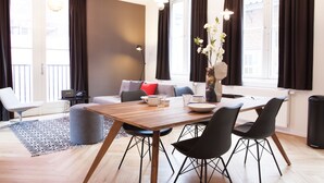 A modern living room with a wooden dining table, black chairs, a grey sofa, and a black floor lamp. - The Duke Apartments ('s-Hertogenbosch)