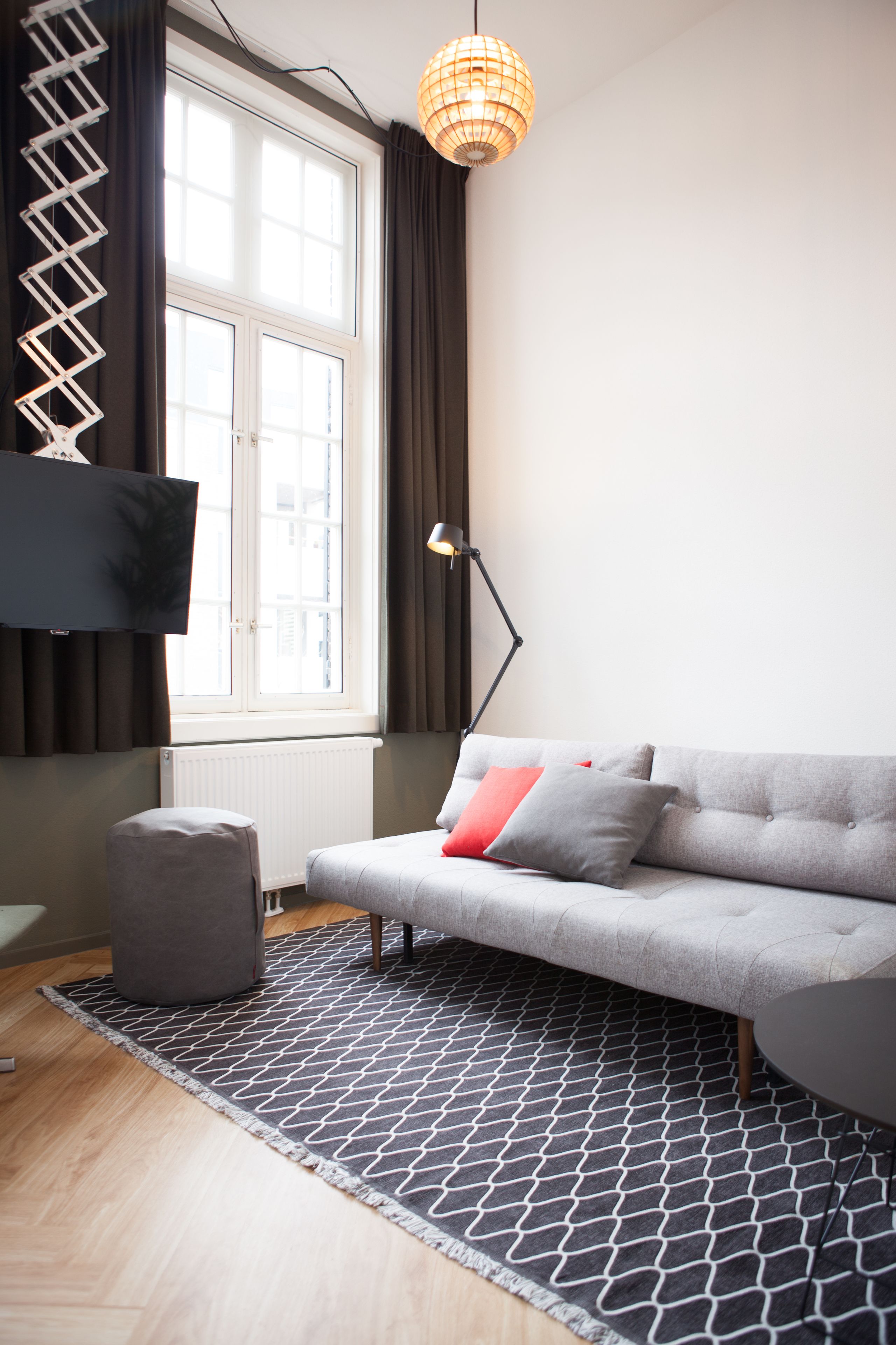 A modern living room with a grey sofa, a black and white patterned rug, a grey ottoman, a floor lamp, and a flat-screen TV mounted on the wall.