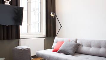 A modern living room with a grey sofa, a black and white patterned rug, a grey ottoman, a floor lamp, and a flat-screen TV mounted on the wall.