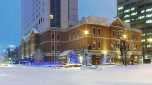 Een hotel met een moderne glazen gevel, een gebouw van baksteen met een traditioneel ontwerp, en een met sneeuw bedekte straat met feestverlichting.