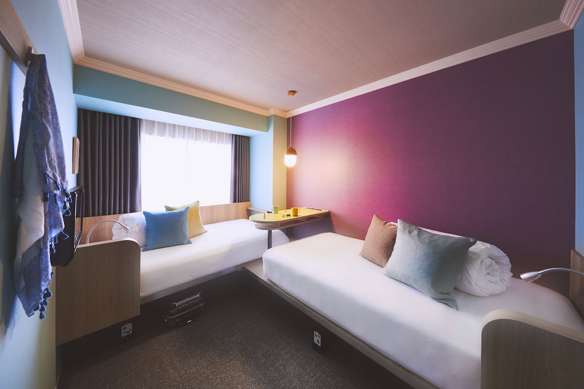 A compact hotel room with two single beds, a small table, and a window with curtains.