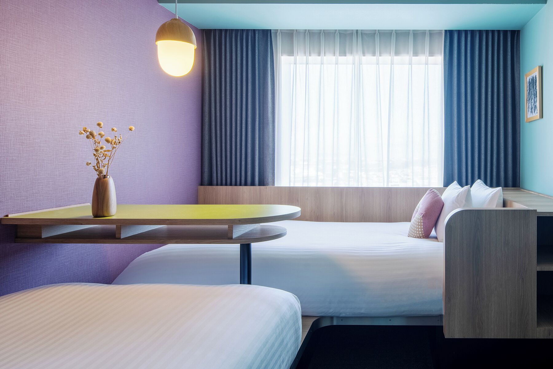 Habitación de hotel con dos camas, una mesita con un florero y una lámpara colgante, además de una ventana con cortinas.