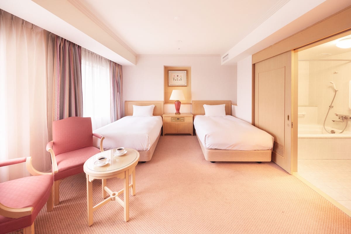 A hotel room with two beds, a chair, a small table, a lamp, and a shower area.