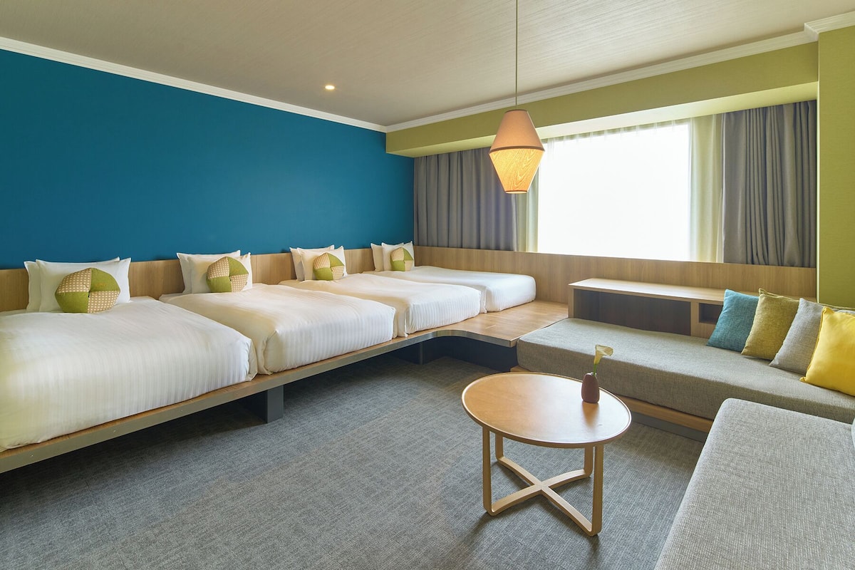 A hotel room with a large bed, a sofa, a small table, and a window with curtains.