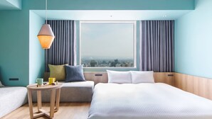Een hotelkamer met een bed, een klein tafeltje, een bank en uitzicht op de stad door grote ramen.