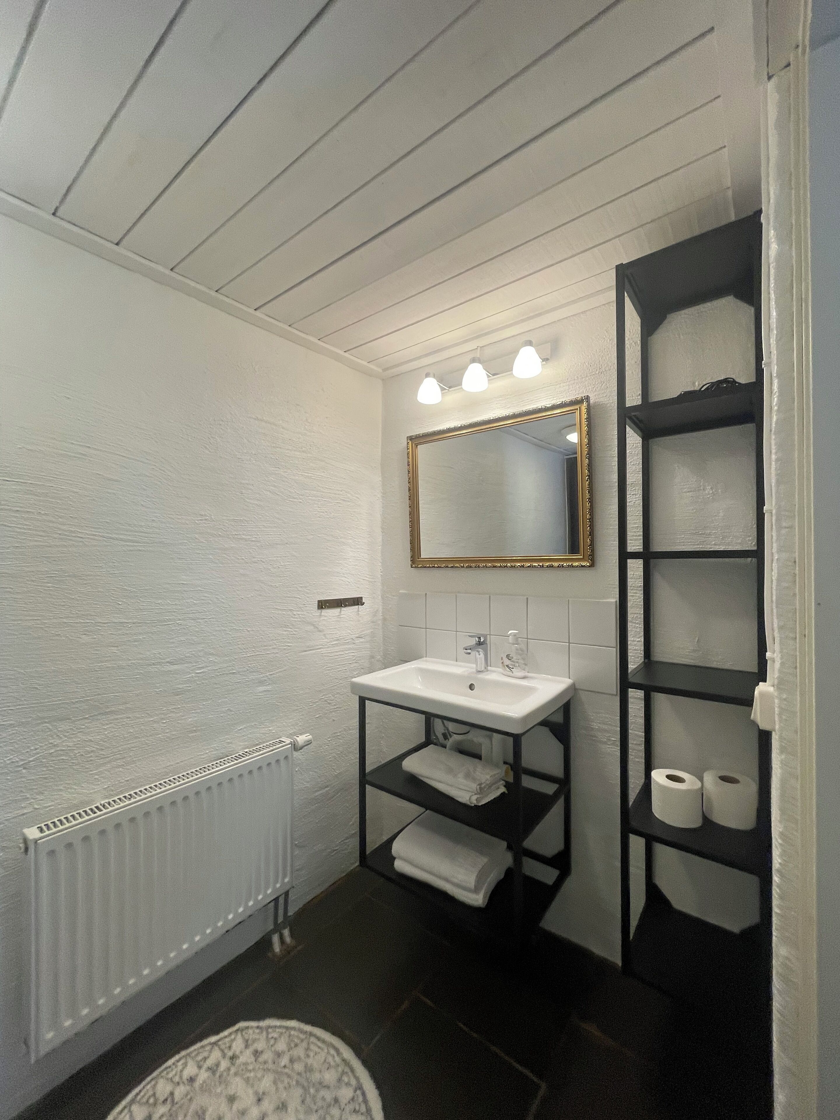 A bathroom with a white sink, a mirror with a gold frame, and a black shelving unit with towels and toilet paper.