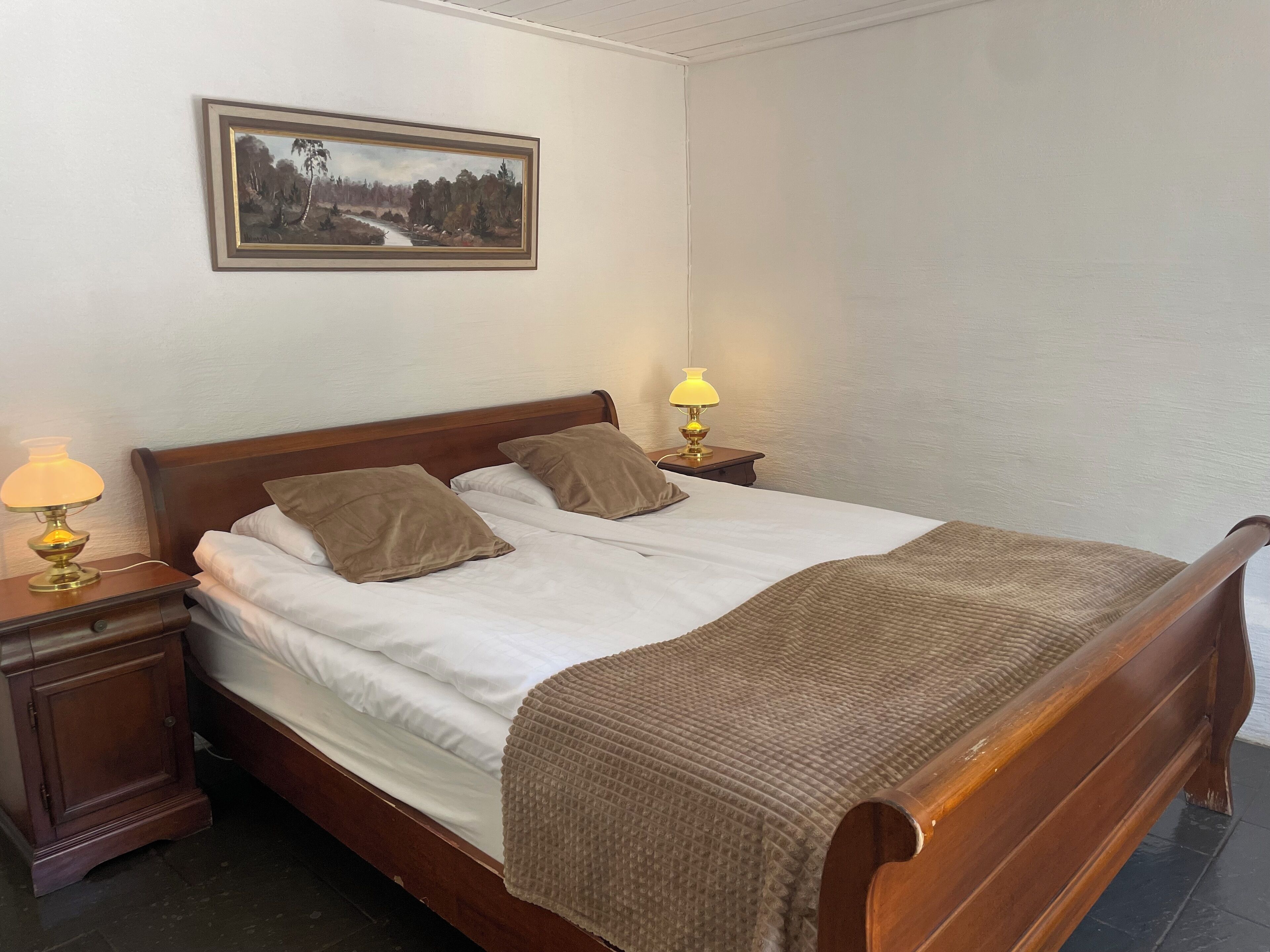 Una cama bien hecha con sábanas blancas y dos almohadas, un cabecero de madera, una mesita de noche con una lámpara y un cuadro con un paisaje en la pared.