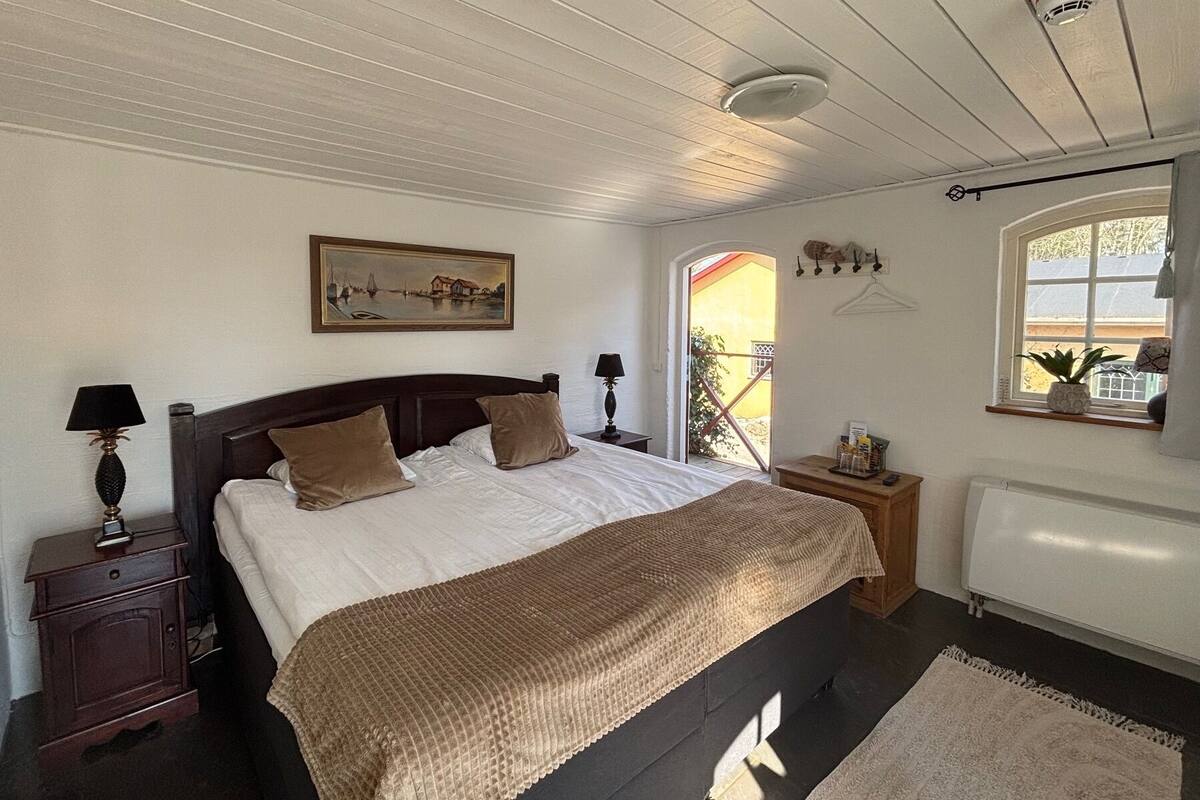 A bedroom with a bed, a nightstand, a lamp, a painting, a window, and a radiator.
