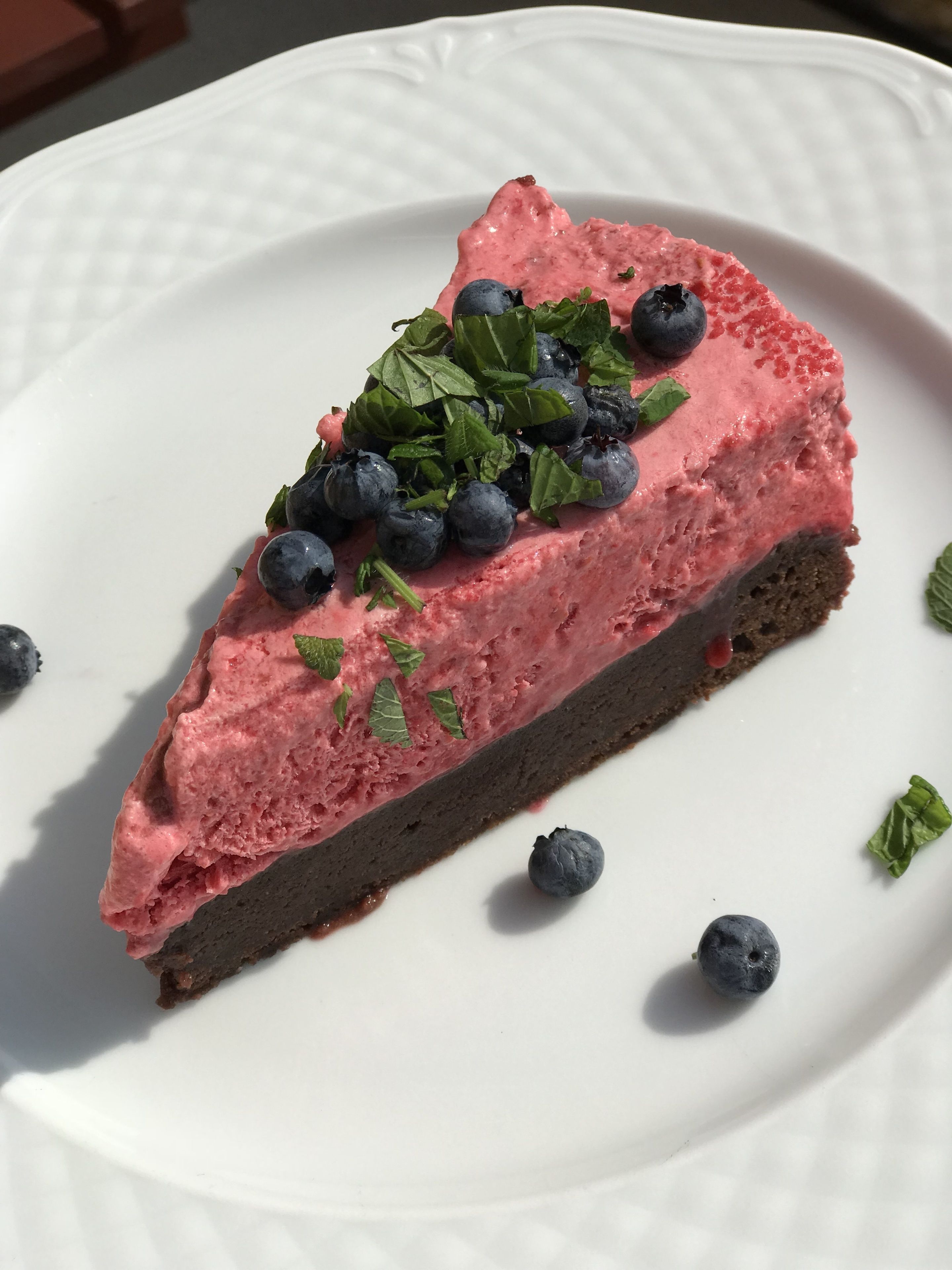 Une part de gâteau au fromage, recouverte d’un glaçage rose et agrémentée de myrtilles, présentée sur une assiette blanche.