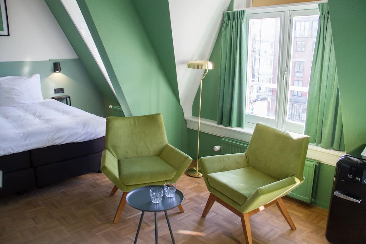 A room with a bed, two green armchairs, a small table with glasses, a floor lamp, and a radiator.