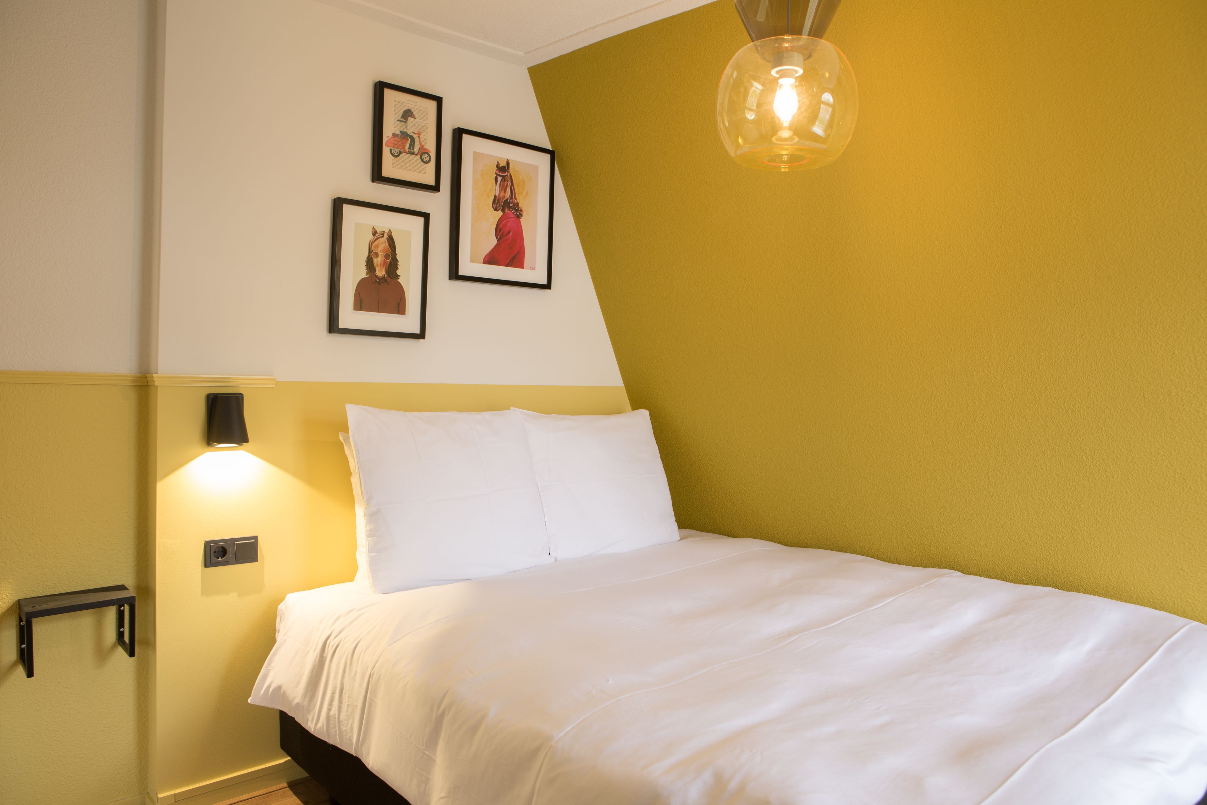 A neatly made bed with white linens in a room with yellow and white walls. There are three framed pictures on the wall above the bed.