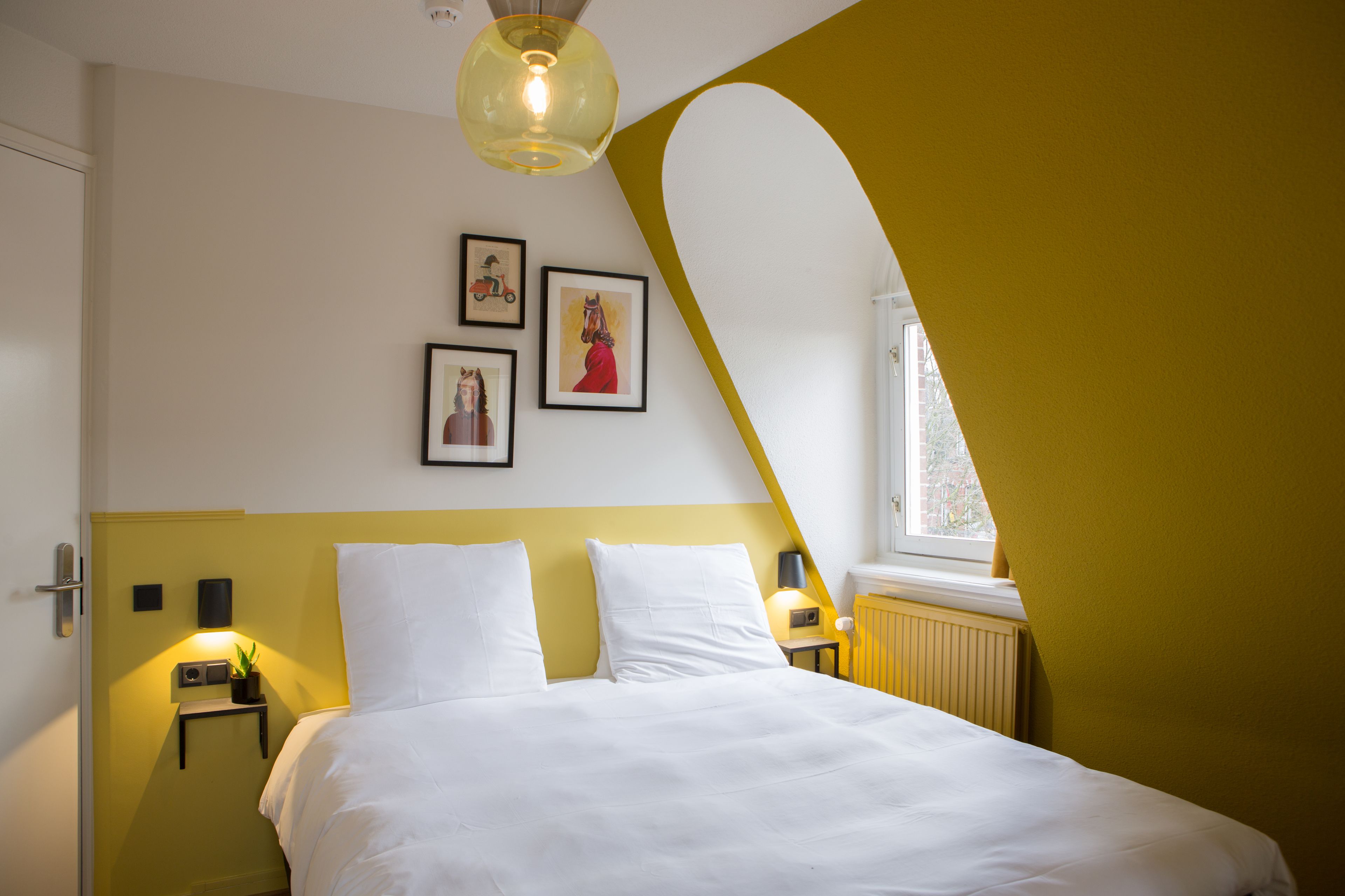 A bedroom with a bed, white bedding, a yellow accent wall, a window, and framed pictures on the wall.