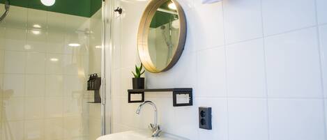 A bathroom with a glass shower enclosure, a white sink with a modern faucet, a round mirror, and a small potted plant on a shelf.