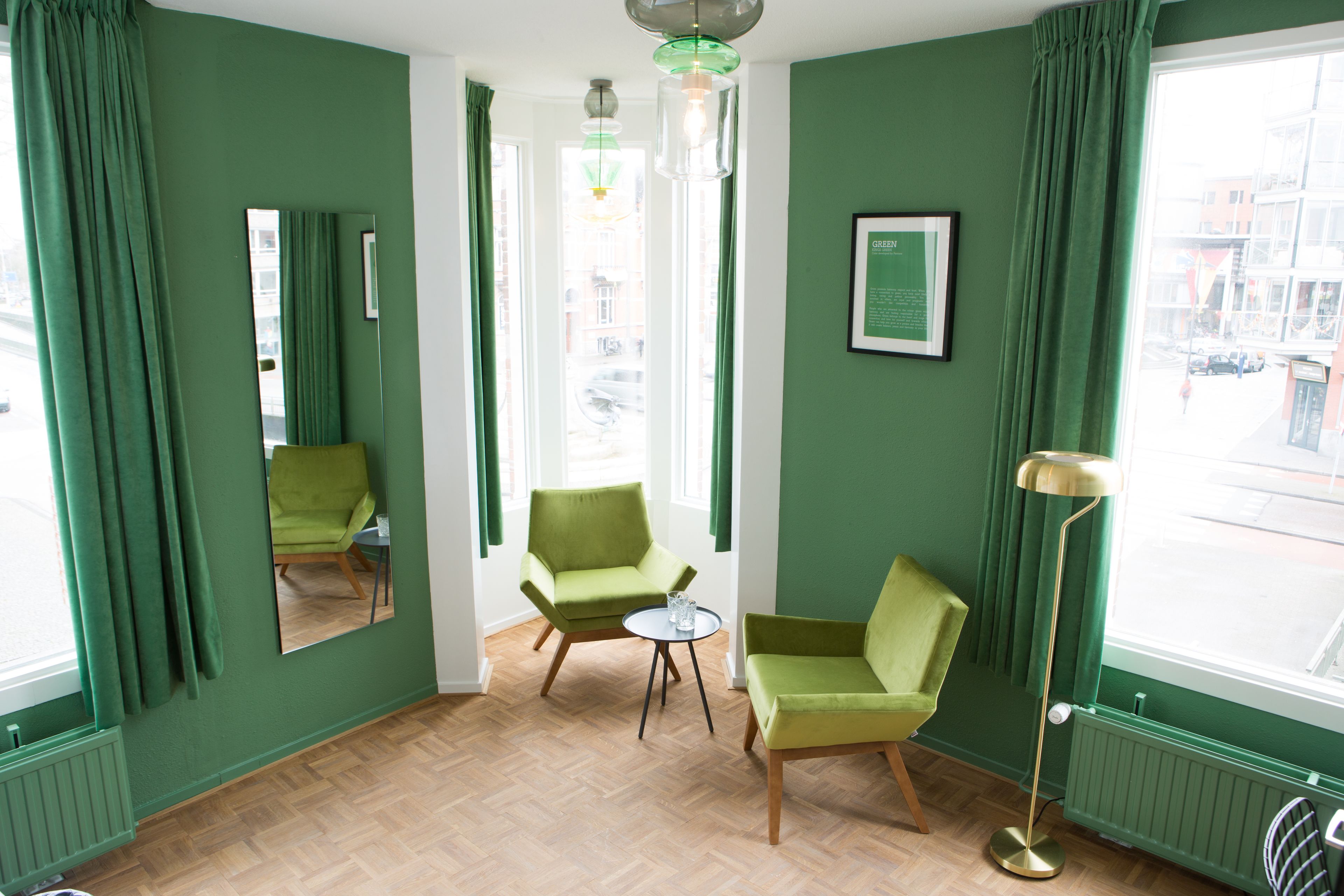 Una habitación con dos sillones verdes, una mesita redonda, una lámpara de pie, un espejo y una ventana que ofrece vistas a edificios.