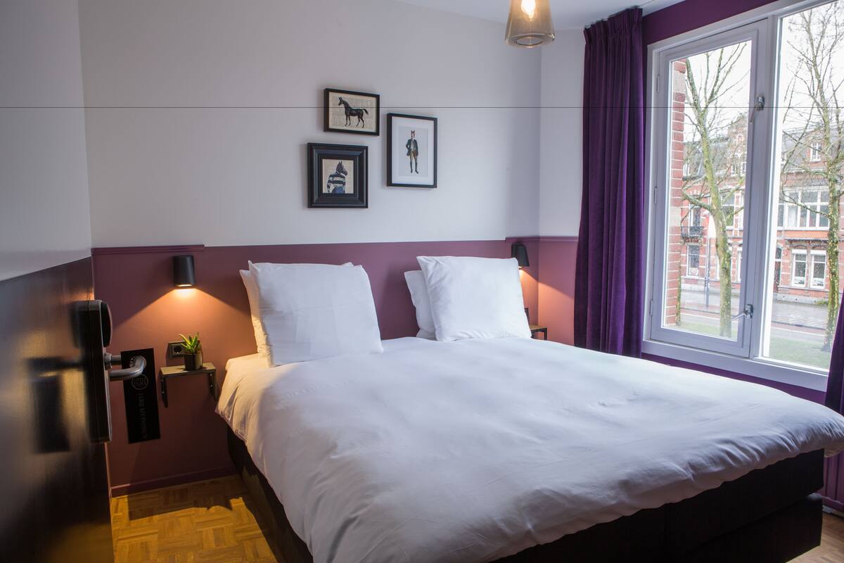 A hotel room with a large bed, a nightstand with a lamp, and a window with curtains.