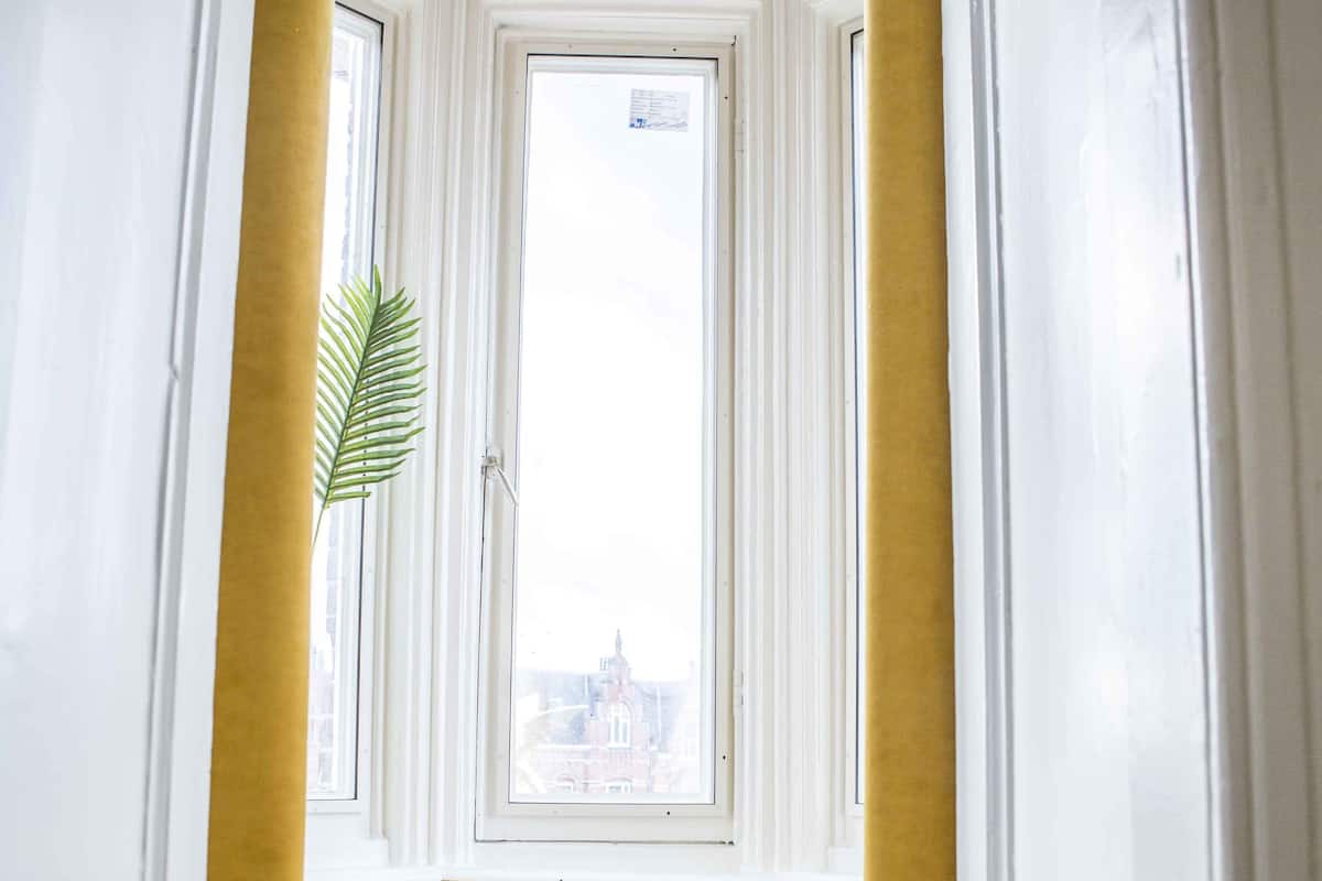 A hallway with a window, a yellow curtain, and a round yellow ottoman.