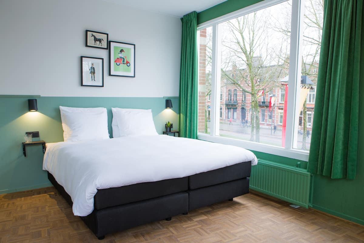 A neatly made bed in a room with a large window, a green wall, and a view of buildings outside.