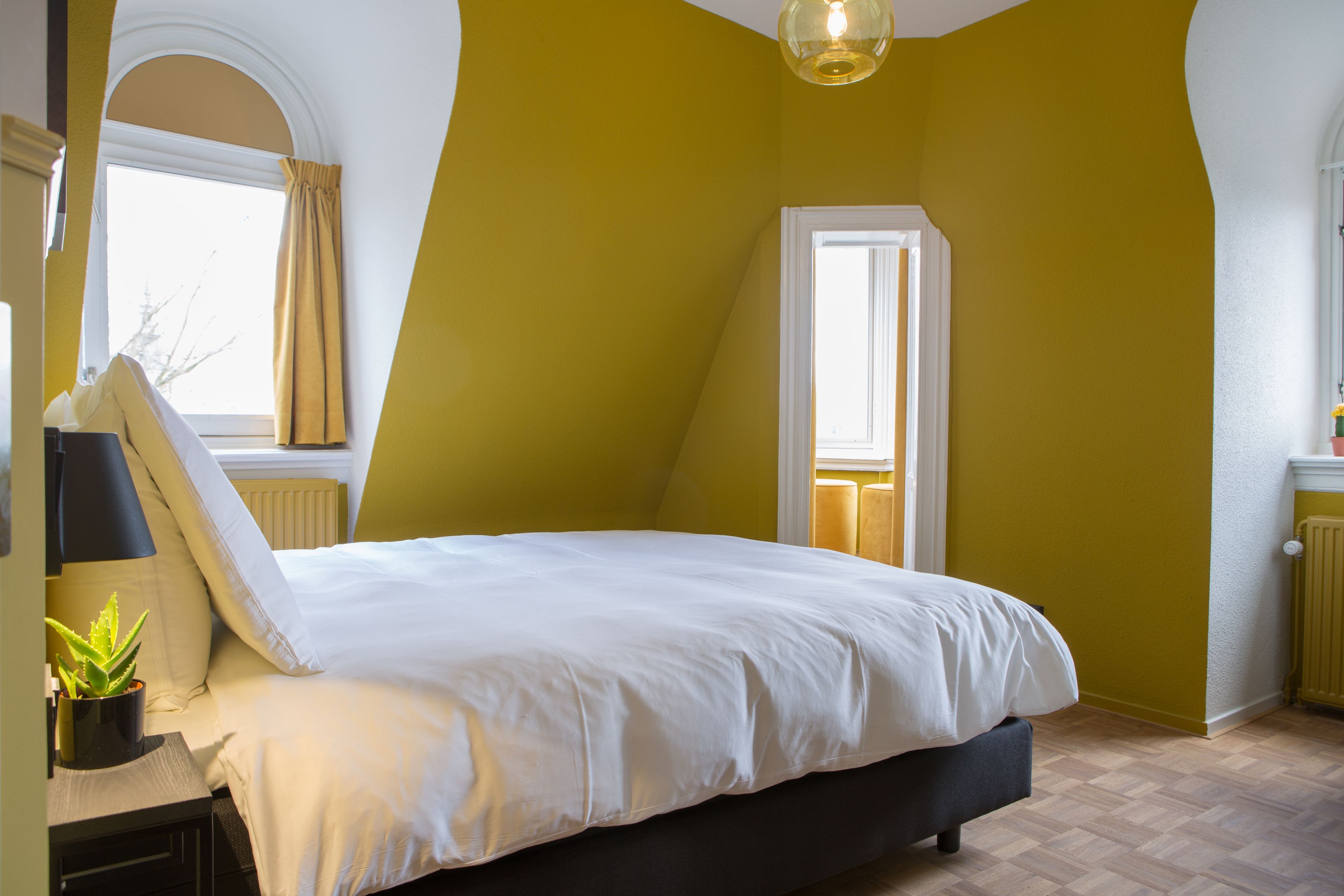 A bedroom with a bed, a nightstand with a lamp, a potted plant, and a window with curtains.