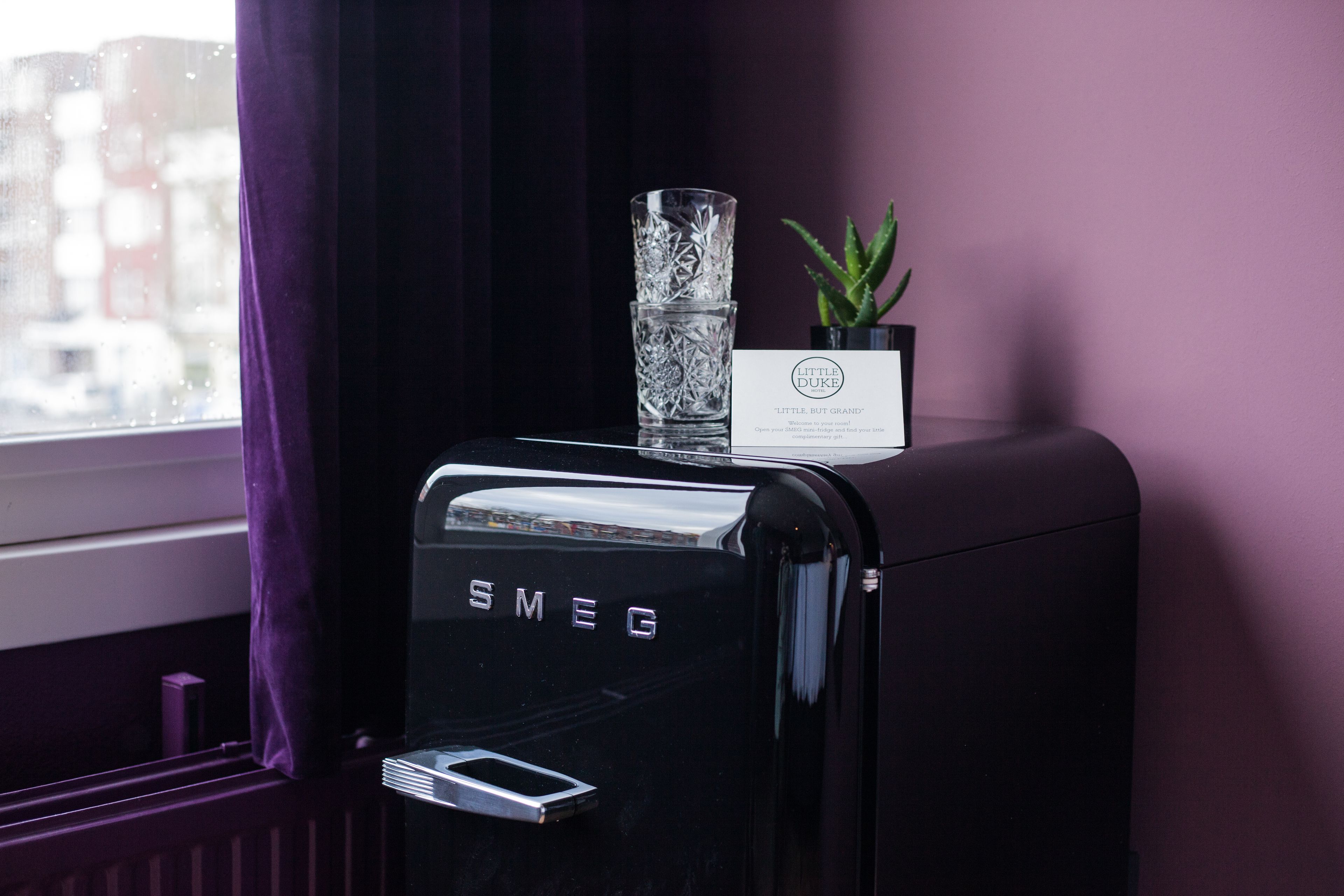 A black Smeg refrigerator with a glass and a card on top, next to a window with purple curtains.