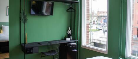 A compact hotel room with a green wall, a bed, a desk with a chair, a TV mounted above the desk, and a window offering a view of the street.