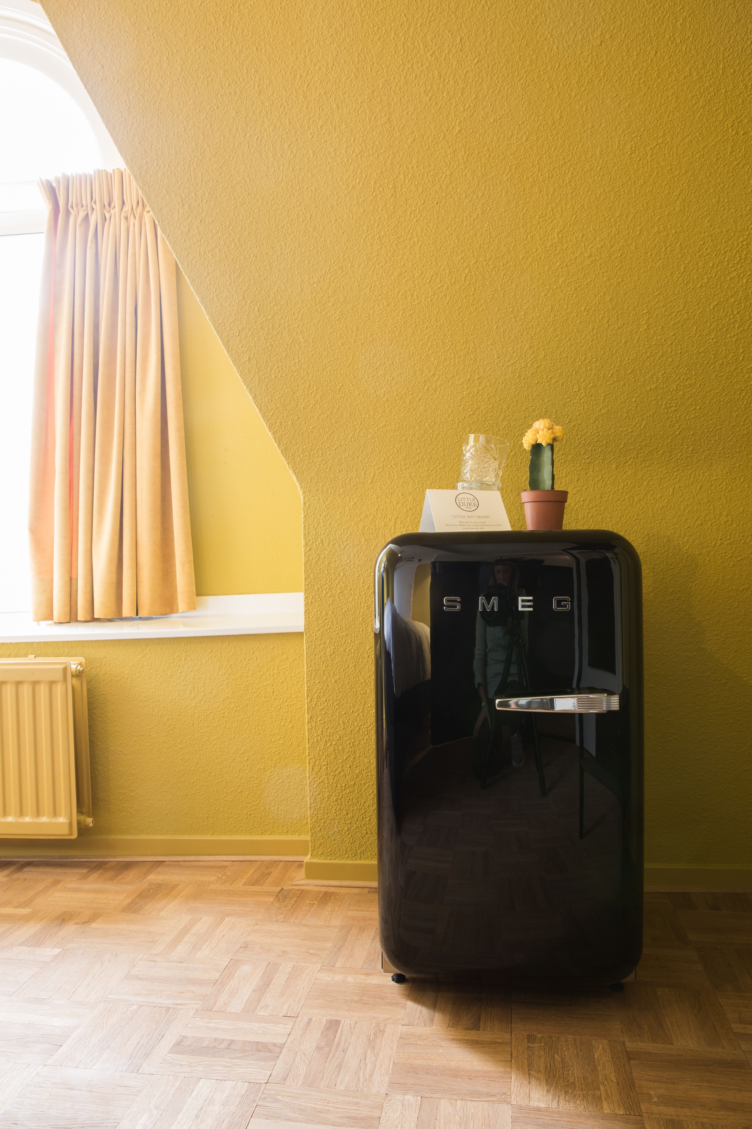 A compact, black suitcase with the brand name 'SMEG' on it, placed on a wooden floor against a yellow wall.