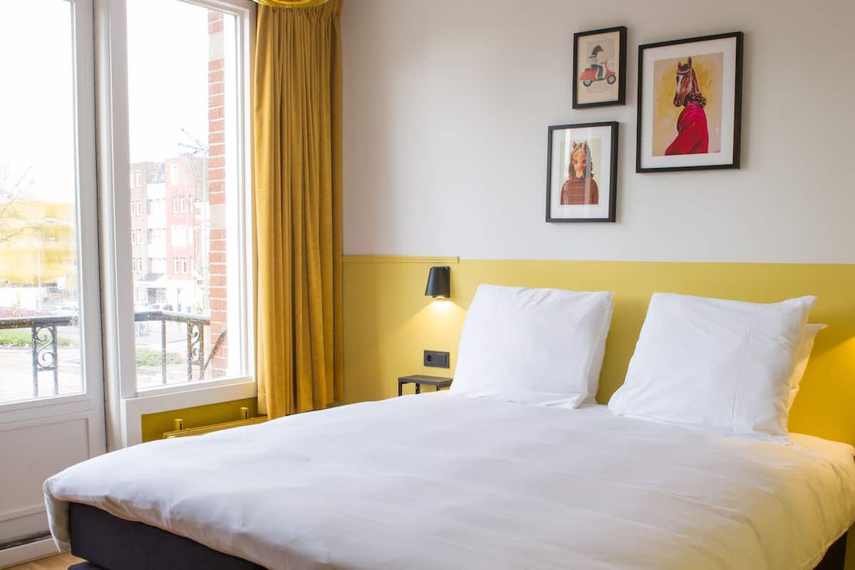 A hotel room with a large bed, a window with curtains, a wall-mounted lamp, and two framed pictures.