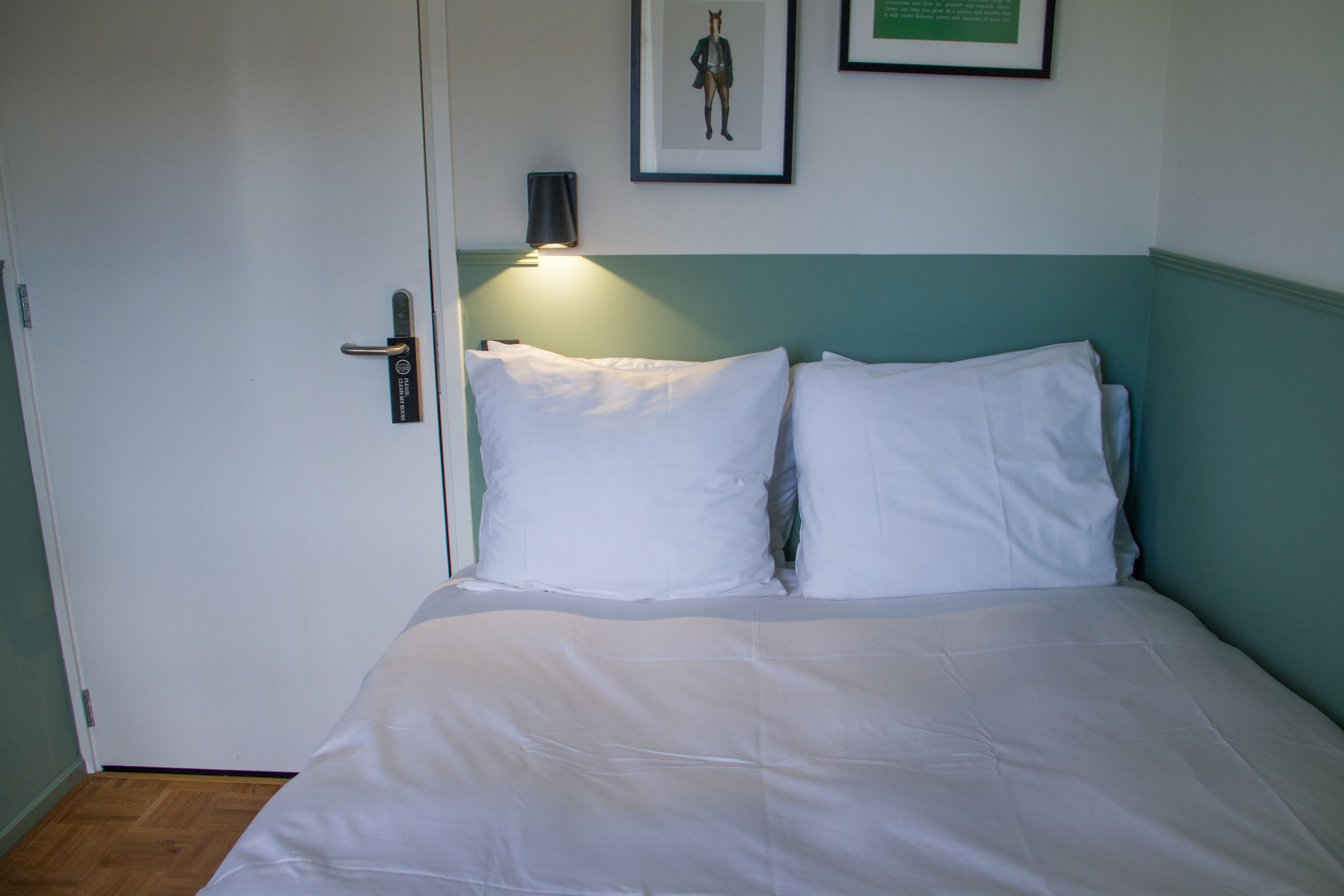 Una cama con ropa de cama blanca, cabecera color aguamarina y lámpara de pared.
