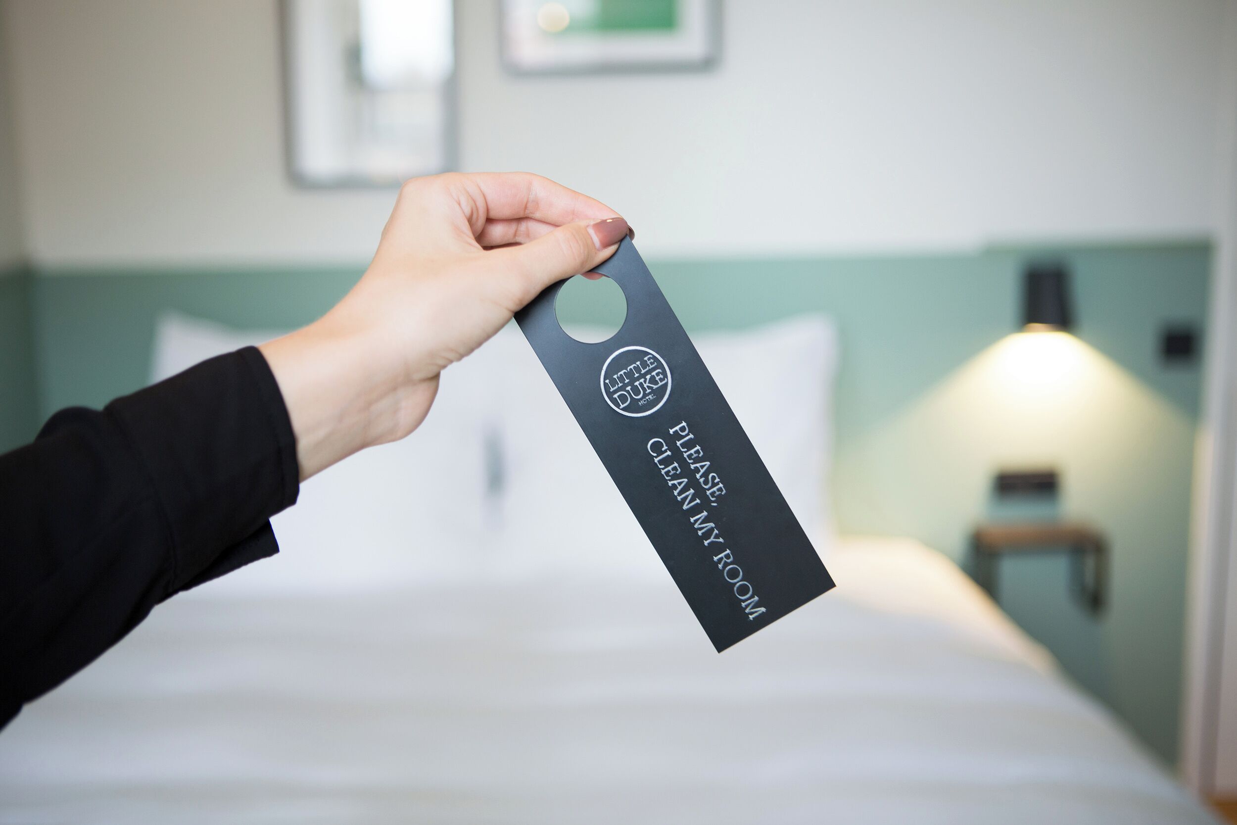 A hand holding a tag with "Little Duke" and "Please clean my room" text, indicating a hotel room service tag.