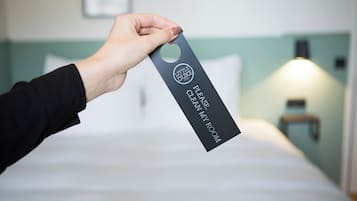 A hand holding a tag with "Little Duke" and "Please clean my room" text, indicating a hotel room service tag.