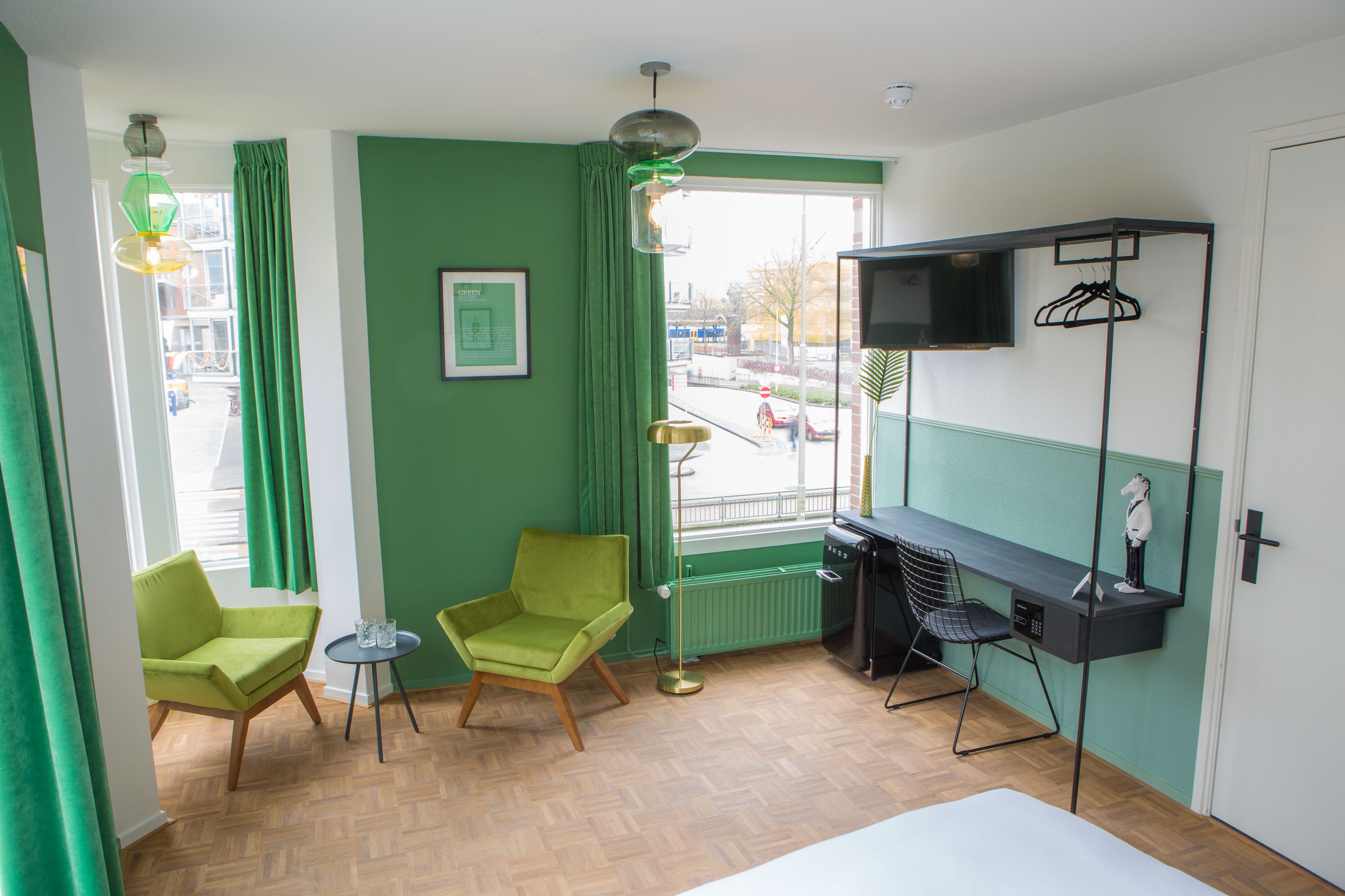 A room with two green chairs, a small table, a bed, a TV on a shelf, and a view of the street outside.
