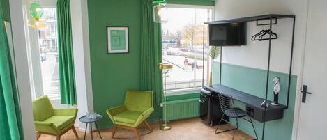 A room with two green chairs, a small table, a bed, a TV on a shelf, and a view of the street outside.