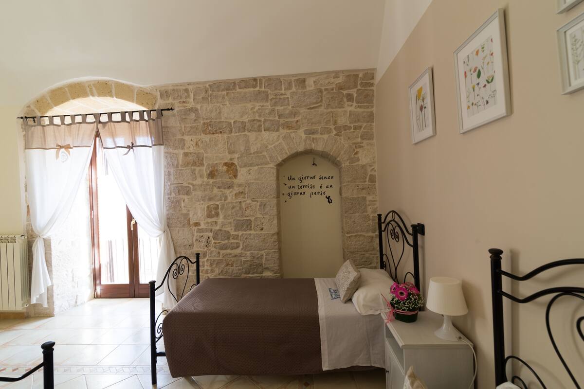A bedroom with a stone wall, a bed with a brown bedspread, a nightstand with a lamp, and a window with curtains.