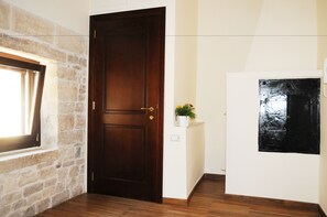 A room with a wooden door, a stone wall, a window with curtains, a small plant on a white shelf, and a black and white artwork on the wall. - B&B di Maria Chiara (Toritto)