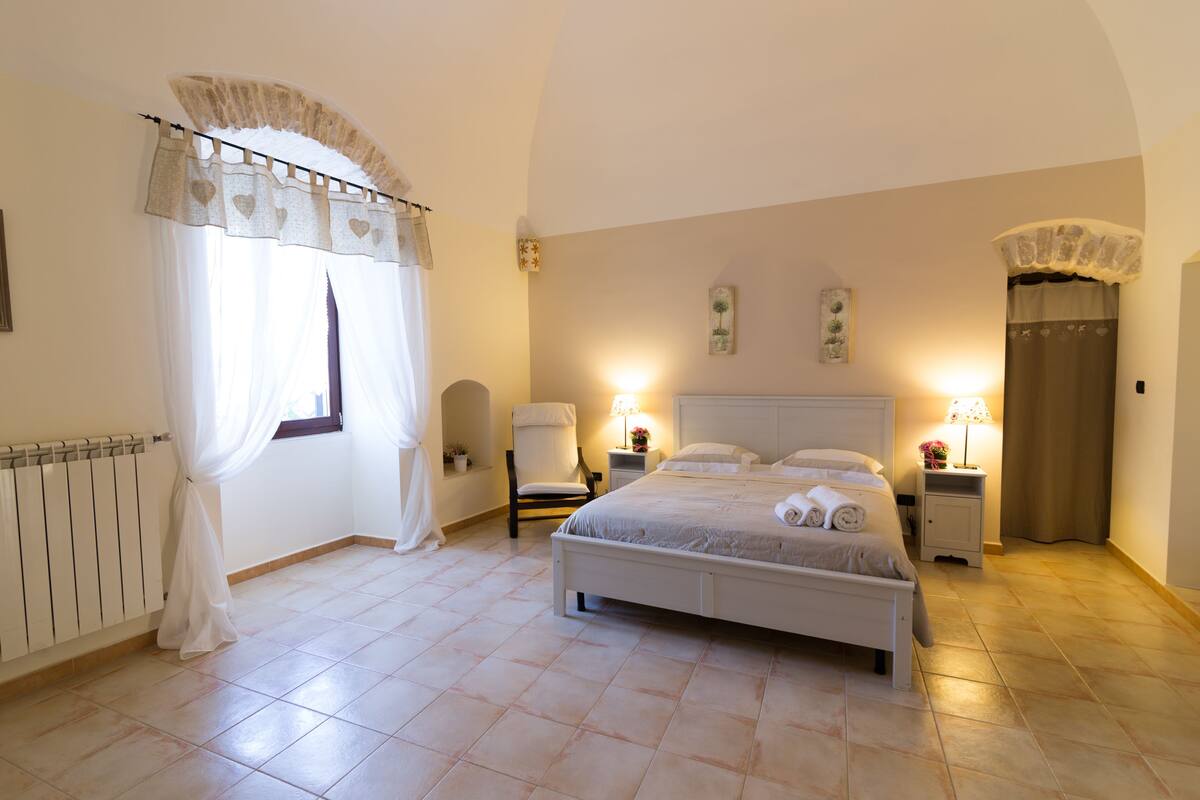 A bedroom with a bed, a chair, a nightstand, a lamp, and a window with curtains.