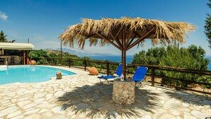 Area piscina con pavimento in pietra, ombrellone di paglia e poltrone da relax blu.