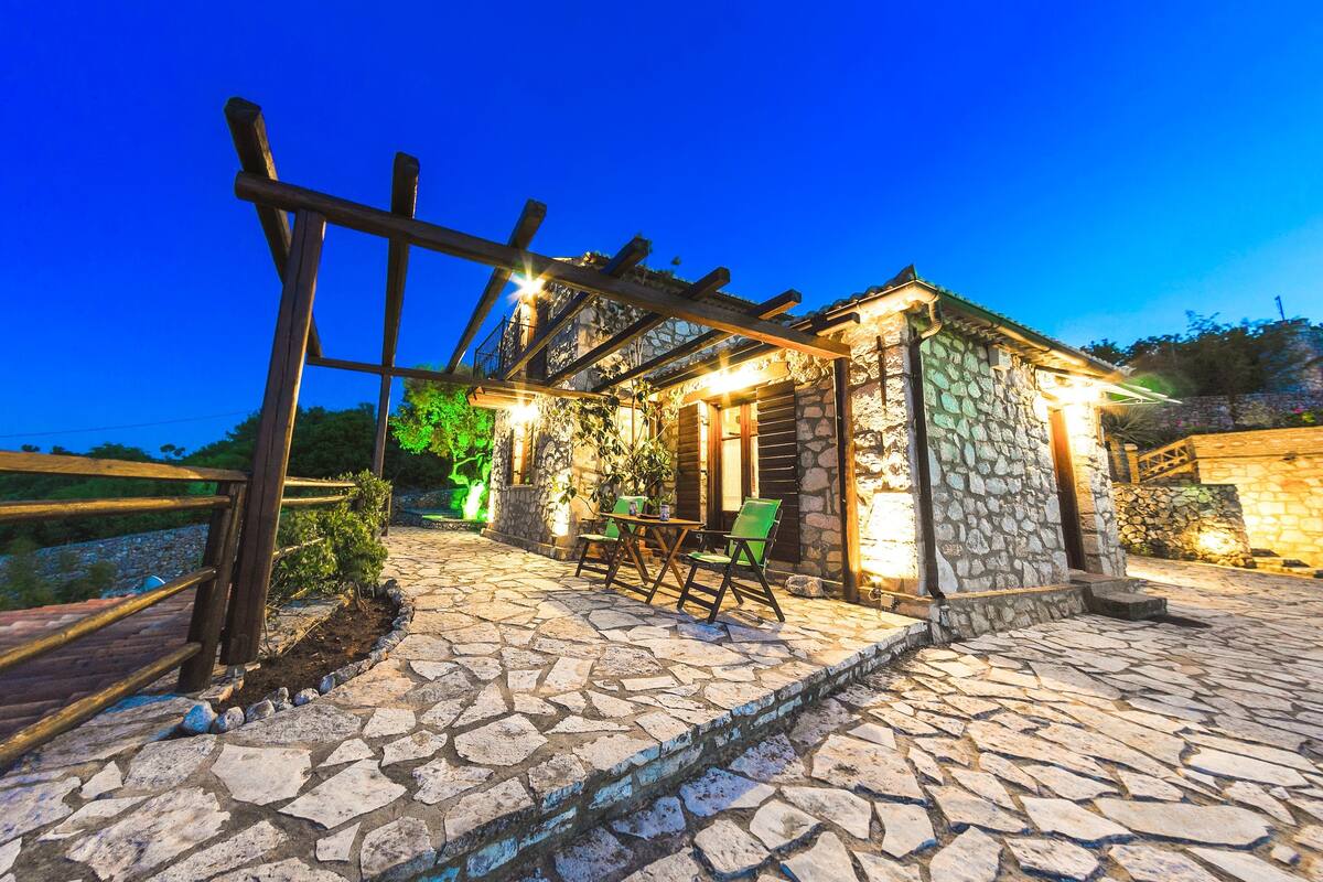 Un edificio de piedra con una pérgola de madera, asientos al aire libre y un sendero de piedra.