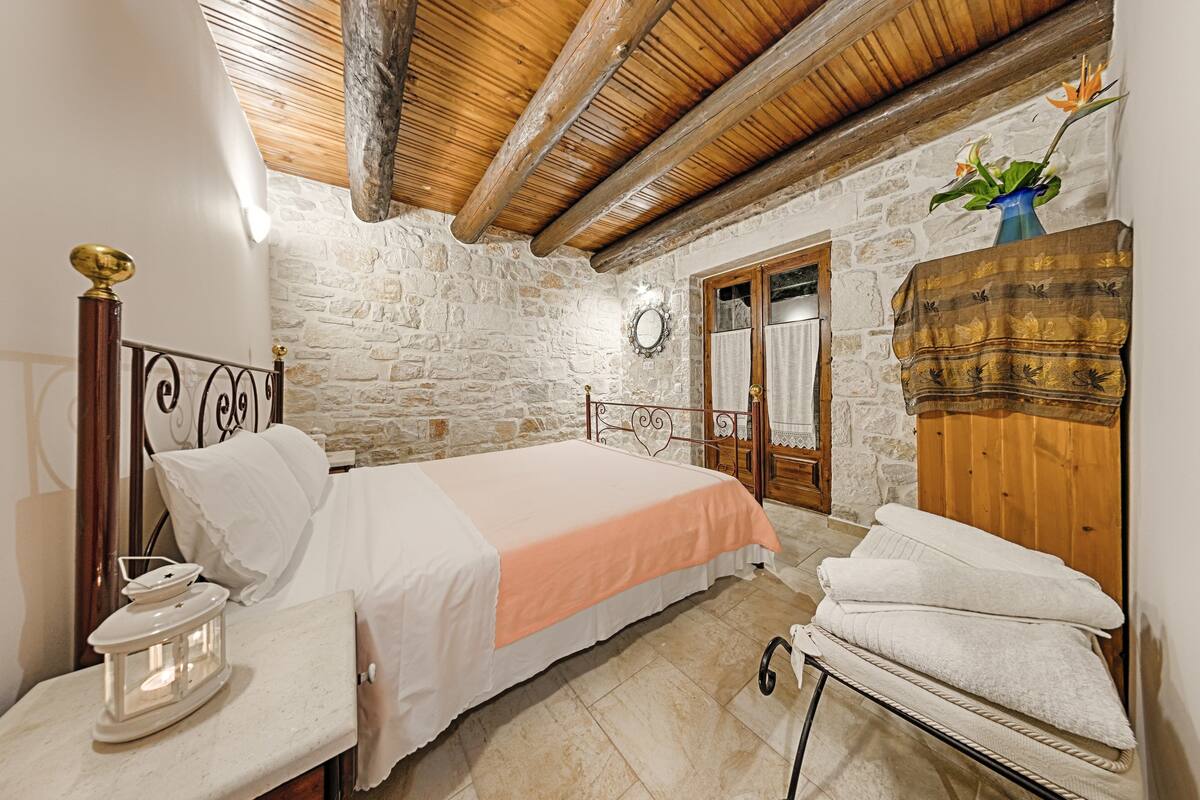 Un dormitorio con pared de piedra, vigas de madera en el techo, una cama con ropa de cama blanca y melocotón, una mesita de noche con lámpara y un armario de madera.