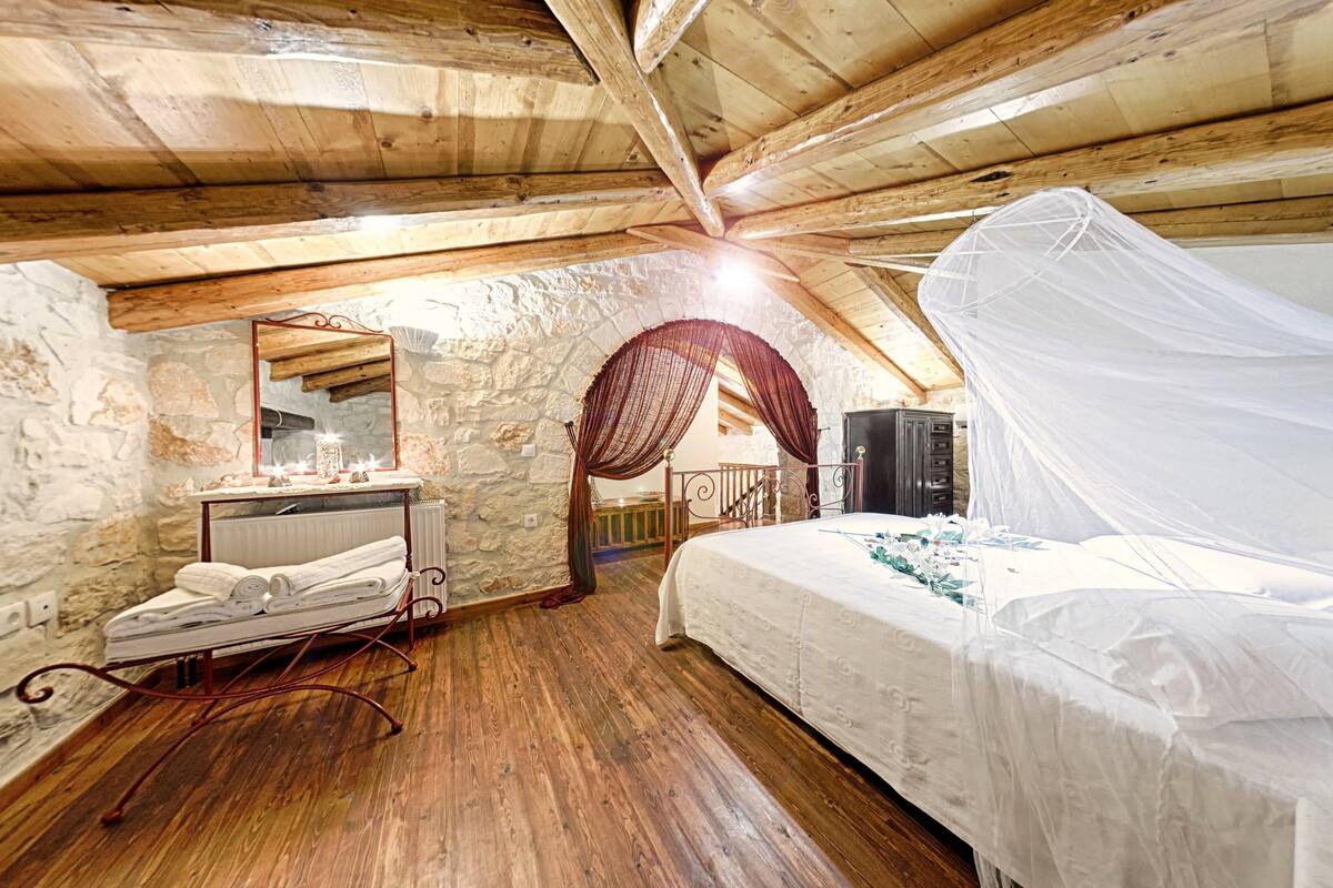 Un dormitorio rústico con pared de piedra, vigas de madera en el techo, una cama con dosel, una mesita de noche con espejo y un radiador.