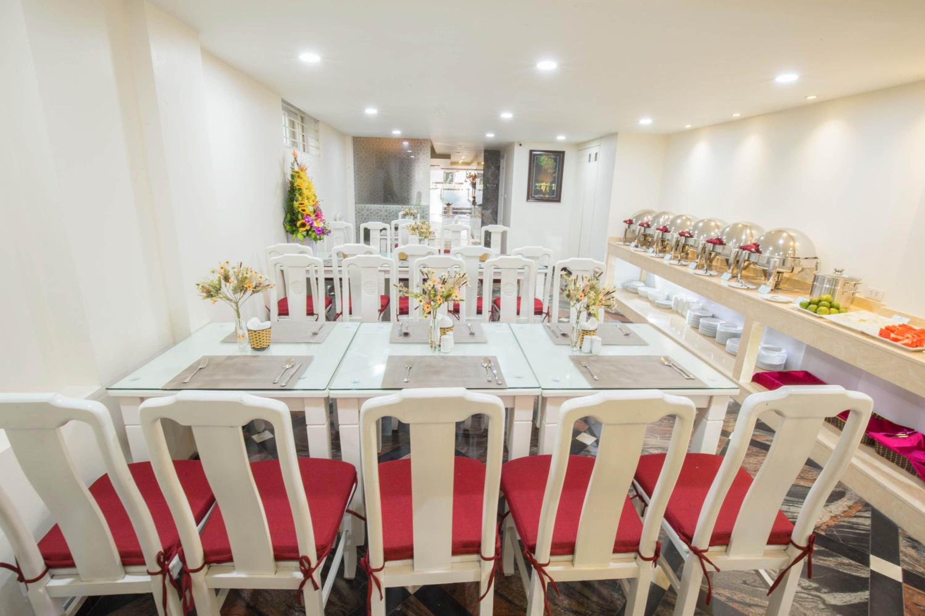 A dining area with white tables and chairs, red cushions, and a buffet line with food and drinks.