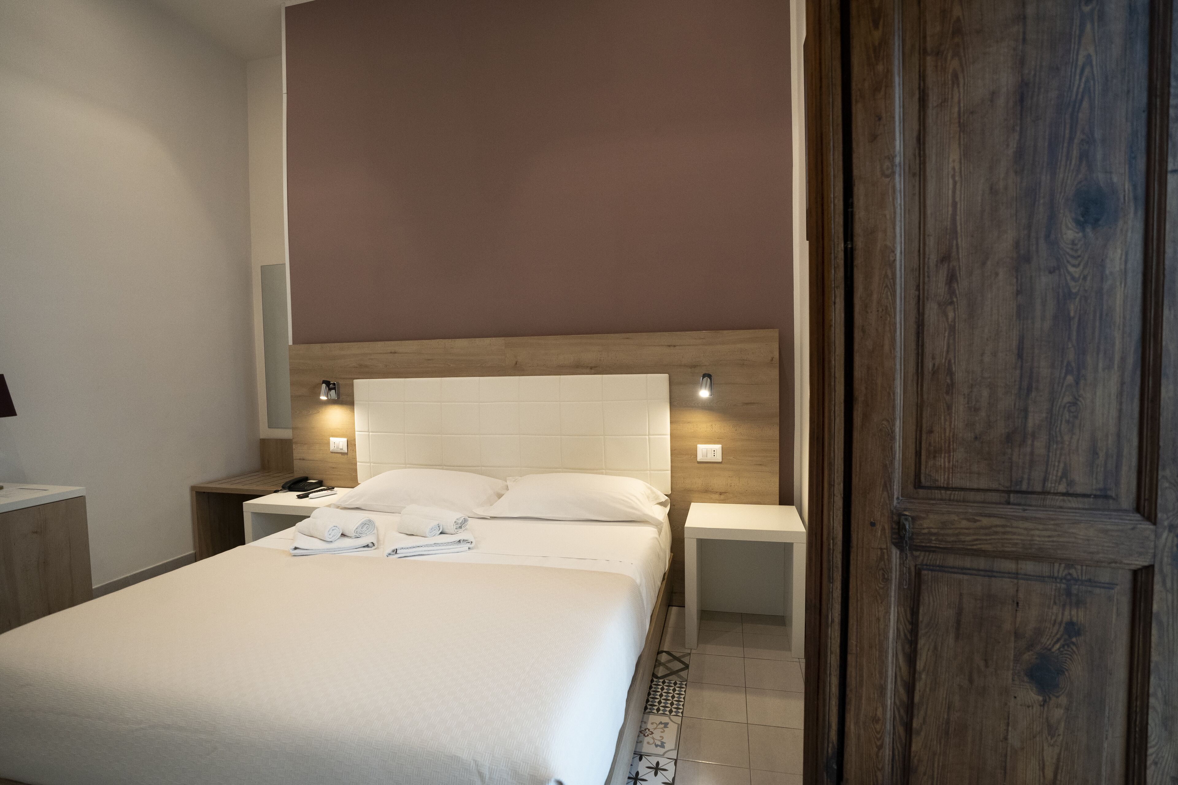 A hotel room with a large bed, two bedside tables, a wooden headboard, and a nightstand with a phone and lamp.