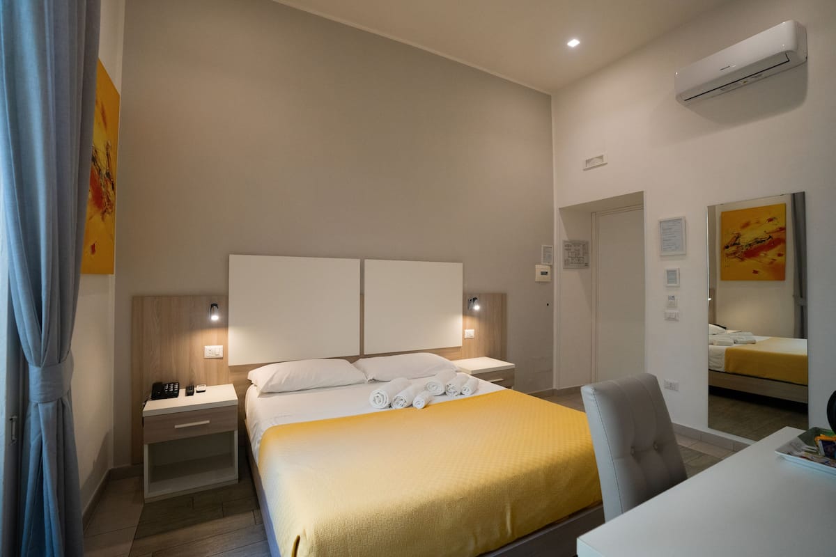 A hotel room with a bed, bedside tables, a desk, and an air conditioning unit.